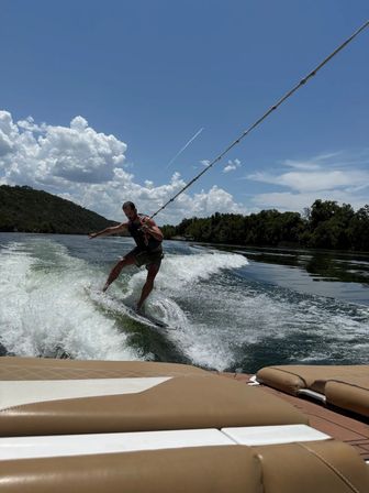 Wakesurfer carving a boat wake on a sunny lake — rider holding the tow rope past tan boat seats, sparkling water, puffy clouds and tree-lined shore under a bright blue sky.