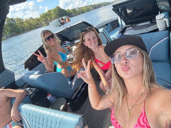 Three friends taking a selfie on a blue-upholstered motorboat during a sunny lake day, two women in bikinis flashing peace signs with a tree-lined shoreline and calm water behind them.