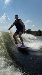 Smiling person wakesurfing on a purple board behind a boat on a sunny lake, carving a green wave with tree-lined shore and blue sky
