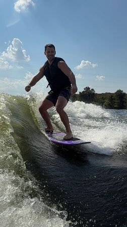 Smiling person wakesurfing on a purple board behind a boat on a sunny lake, carving a green wave with tree-lined shore and blue sky