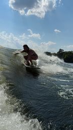 Smiling surfer wakesurfing a boat-generated wave on a sunny lake, crouched on the board with splashing water, blue sky, puffy clouds and tree-lined shore in the background.