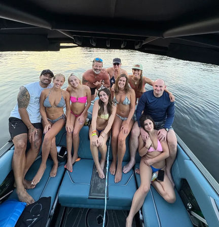 Group of ten people in swimsuits smiling and posing on a blue motorboat under a black canopy, docked on a calm lake with soft sunset light in the background.