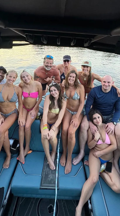 Group of ten people in swimsuits smiling and posing on a blue motorboat under a black canopy, docked on a calm lake with soft sunset light in the background.