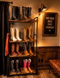 Rustic western boutique interior with wooden shelves displaying multiple cowboy boots (black, white, tan, pink), lit by a wall sconce next to a framed sign reading "PUT YOUR BEST FOOT FORWARD", leather bench and wood-paneled floor.