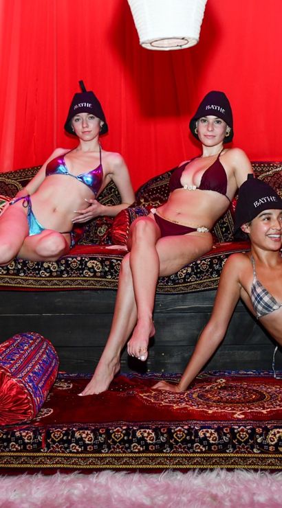 Three women in bikinis and black bucket hats lounging on a low platform inside a red cabana decorated with patterned Moroccan-style rugs, bolsters and cushions.