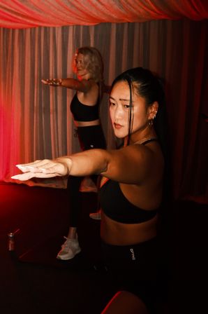 Two women in a red-lit indoor studio holding arms forward in a focused yoga/fitness pose, wearing black workout outfits in front of draped curtains.