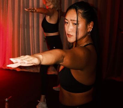 Two women in a red-lit indoor studio holding arms forward in a focused yoga/fitness pose, wearing black workout outfits in front of draped curtains.