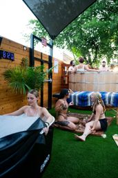 Outdoor spa patio scene with friends in swimsuits—people chatting in a wooden hot tub and sitting on a rug on artificial turf under a shade sail and leafy trees, relaxed summer vibe.