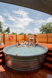 Five people relaxing and sipping drinks in a round galvanized hot tub on a sunny wooden deck with a life preserver on the fence and blue sky overhead