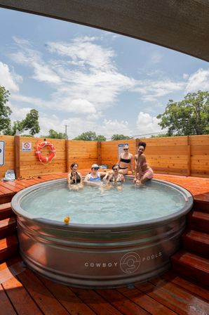 Five people relaxing and sipping drinks in a round galvanized hot tub on a sunny wooden deck with a life preserver on the fence and blue sky overhead