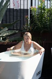 Person in a white swimsuit relaxing in an outdoor soaking tub on a sunny patio, hands on the tub edges with a visible tattooed arm, surrounded by tall green plants and a decorative paper lantern.