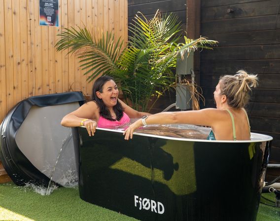 Two women laughing in a black outdoor cold-plunge tub on a sunny wooden-fenced patio with palm plants and artificial turf.
