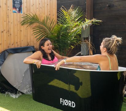 Two women laughing in a black outdoor cold-plunge tub on a sunny wooden-fenced patio with palm plants and artificial turf.