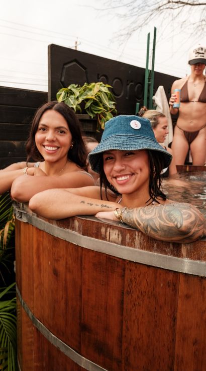 Two smiling women leaning on the rim of a wooden outdoor hot tub — one wearing a denim bucket hat with a tattooed arm — others in swimwear and potted plants in the background.