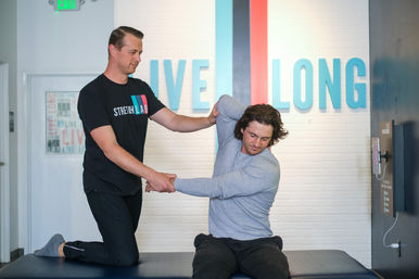 Trainer in a black shirt gently assists a seated man in a gray long-sleeve with a shoulder and torso stretch on a padded table inside a bright fitness studio with a white brick wall and colorful wall lettering.
