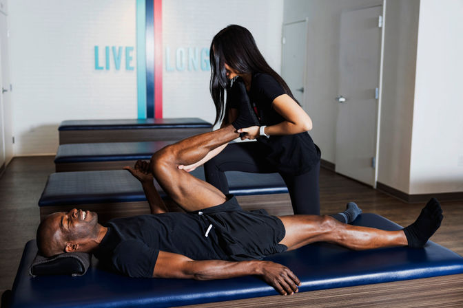 Smiling man lies on a blue therapy table while a female therapist stretches his raised leg in a modern physical therapy/fitness studio with a colorful 'LIVE LONG' wall sign