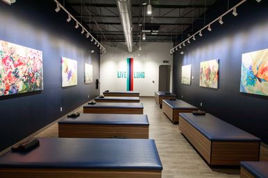 Modern wellness studio interior with rows of blue-padded treatment tables, colorful abstract paintings on dark walls, and a 'LIVE LONG' mural on a white brick end wall.