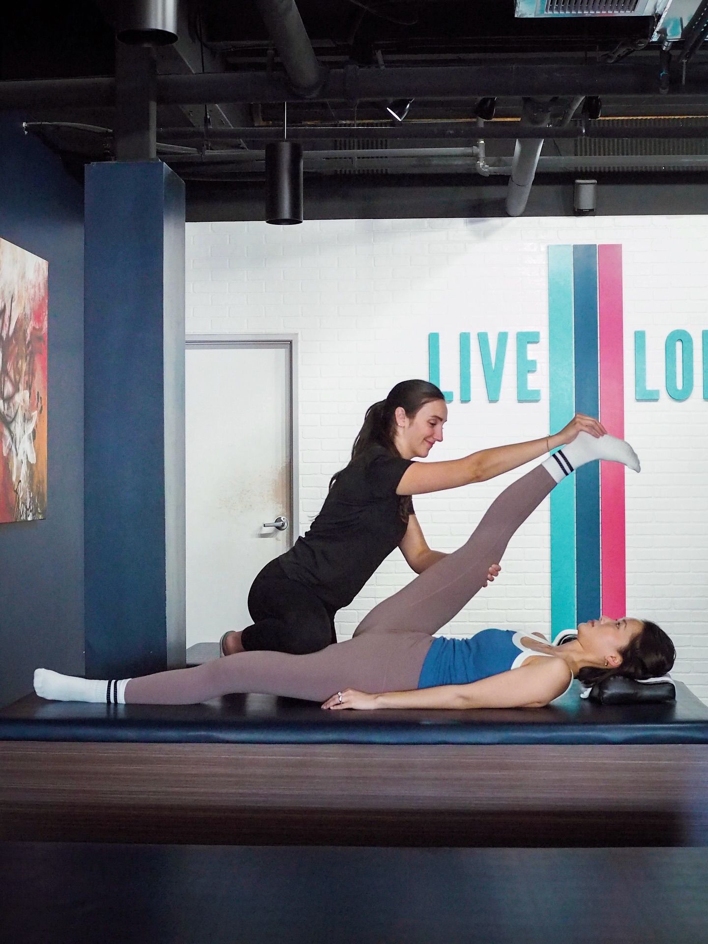 Physical therapist helping a woman perform a supine hamstring stretch on a treatment table in a modern wellness studio with colorful vertical stripes and 'LIVE' lettering on a white brick wall.