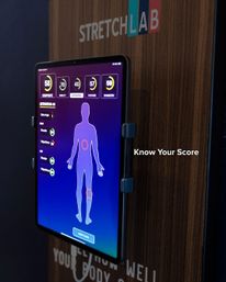 Tablet-mounted interactive fitness kiosk on a wood panel showing a digital body silhouette with highlighted joints, mobility and posture scores, and the prompt 'Know Your Score'.