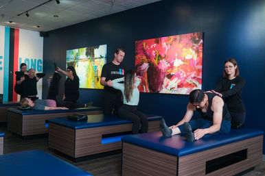 Group assisted stretching session in a modern fitness studio, trainers helping clients on blue padded tables against navy walls with vibrant abstract paintings.