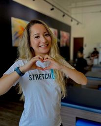 Smiling fitness instructor forming a heart with her hands in a modern stretch studio with padded stretch tables and colorful wall art