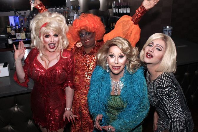 Four drag performers in vibrant wigs and sequined costumes posing and smiling at a bar counter during a lively drag show, colorful outfits and bottles visible in the background.