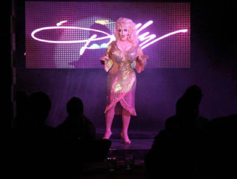 Drag performer in a sparkling pink sequin dress and blonde wig on a purple-lit stage with silhouetted audience watching a cabaret-style show