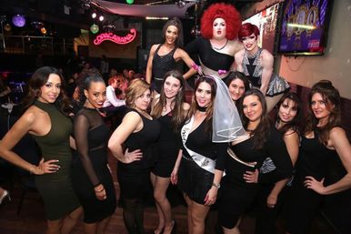 Bachelorette party group of women and drag performers posing in a lively nightclub with colorful stage lights and disco balls; bride-to-be wearing veil and sash.