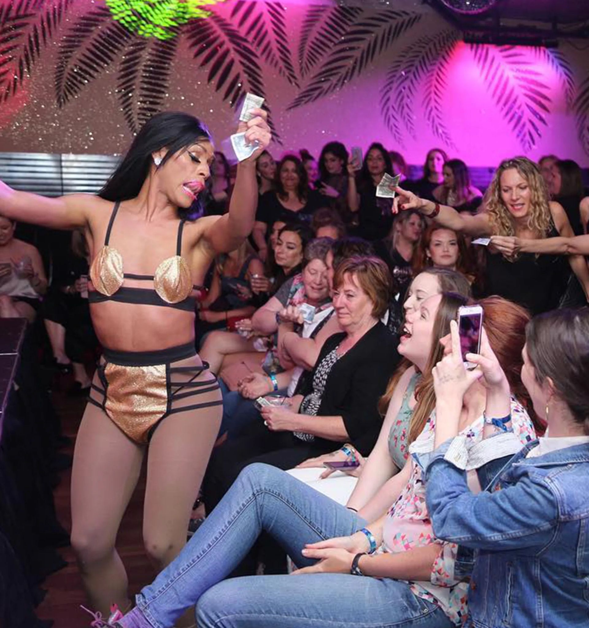 Drag performer in gold costume dancing at a pink-lit nightclub, crowd of women cheering and handing cash near a tropical palm-patterned wall.