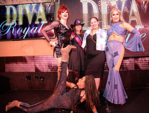 Five people pose under a large "DIVA Royal" stage sign at a colorful drag show—two glittery drag performers, two smiling guests with sashes, and a performer doing a dramatic floor split.