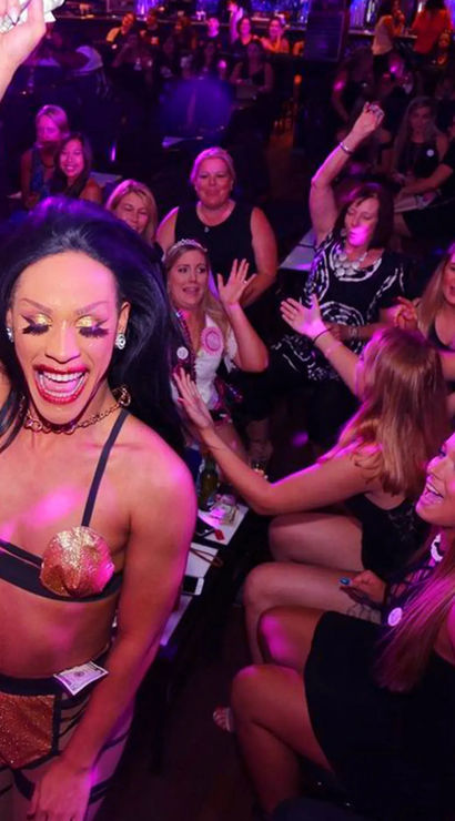 Energetic drag performer in glittery gold costume raising an arm on stage while a cheering crowd of women enjoy a bachelorette-style night out in a purple-lit nightclub