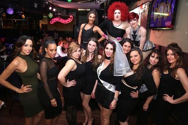 Bachelorette party group photo in a lively nightclub: bride-to-be with veil and sash surrounded by friends in black dresses and colorful drag performers under disco lights and neon signage