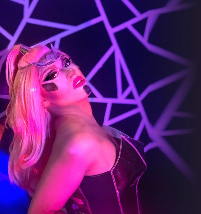 Glamorous drag performer in pink and purple neon lighting, wearing a long blonde wig, dramatic makeup and a futuristic black corset, posing against a geometric neon backdrop on a nightclub stage