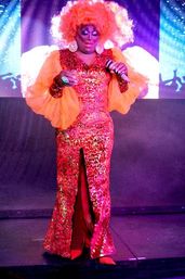 Drag performer in a sparkling red-and-gold sequined gown and voluminous orange wig holds a microphone on a colorful nightclub stage
