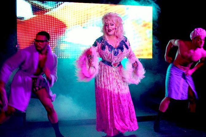 Drag performer in a glamorous pink gown with feathered sleeves and a big blonde wig center stage, flanked by two male backing dancers, colorful LED backdrop and stage fog at a lively nightclub drag show.