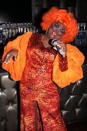 Colorful drag performer in a fluffy orange wig and voluminous sleeves, wearing a glittery red-orange gown and holding a rhinestone microphone in a dim nightclub bar.