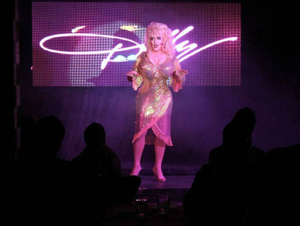 Glamorous drag performer in a sparkling pink sequin dress and blonde wig on a cabaret stage with neon LED backdrop and silhouetted audience.