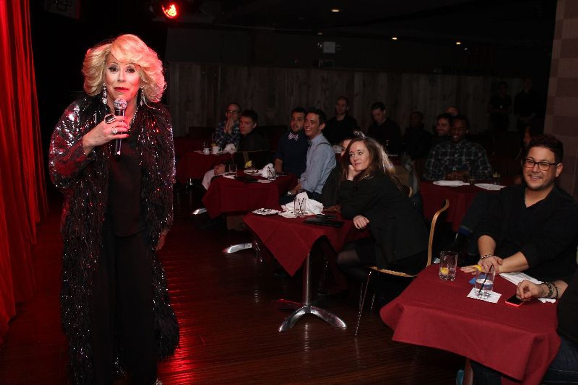 Blonde performer in a sparkly black coat holding a microphone on stage at an intimate cabaret-style comedy club, patrons seated at red-tablecloth dinner tables watching and smiling.