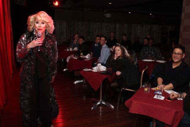 Blonde performer in a sparkly black coat holding a microphone on stage at an intimate cabaret-style comedy club, patrons seated at red-tablecloth dinner tables watching and smiling.