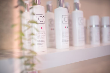Spa skincare shelf with white bottles, front labeled Lavender Cleansing Crème, soft pink glow and blurred trailing greenery