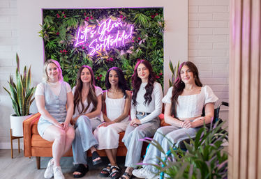 Five women seated on an orange sofa in a bright studio lounge with a lush vertical plant wall, pink neon sign, potted plants, and a relaxed modern vibe.
