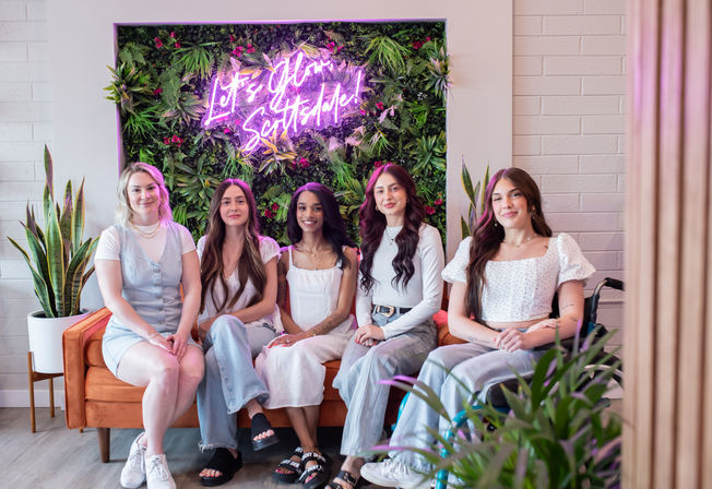 Five women seated on an orange sofa in a bright studio lounge with a lush vertical plant wall, pink neon sign, potted plants, and a relaxed modern vibe.