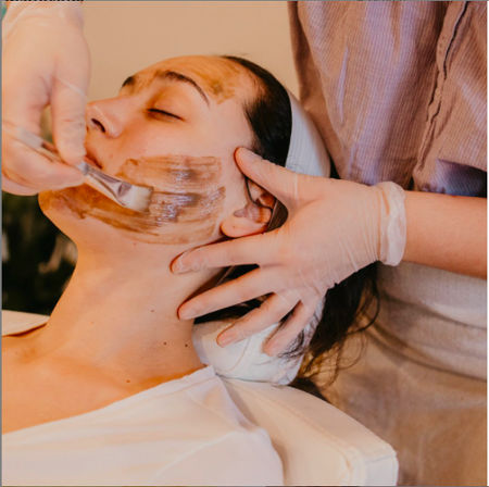 Close-up of a relaxed client receiving a spa facial as a skincare professional in gloves spreads a brown mask across the cheek with a spatula while hair is held back with a headband.