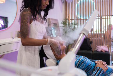 Esthetician in a white dress using a facial steamer over a client reclined in a spa chair during a skincare treatment, modern salon interior with neon mirror and wooden slat wall.