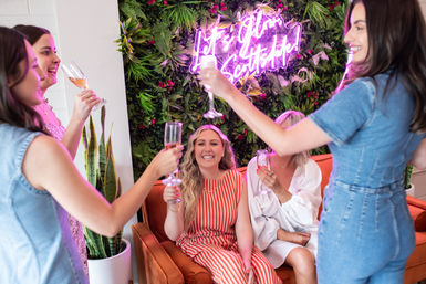 Women toasting with champagne on an orange sofa in a trendy indoor lounge with a neon sign and lush plant wall.