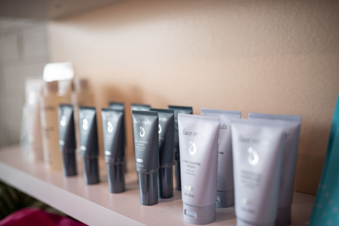 Neatly lined facial skincare tubes and creams on a light-colored shelf in a softly lit spa or salon, gray and lavender packaging on display.
