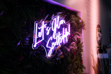 Purple-pink neon sign reading "It's Your Scalp" mounted on a lush living wall of tropical plants, casting a vibrant glow across a modern salon or spa interior.
