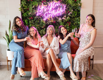 Five smiling women toasting with champagne on an orange couch in front of a lush green wall and neon “Let’s Glow Scottsdale” sign, indoor celebration vibe.