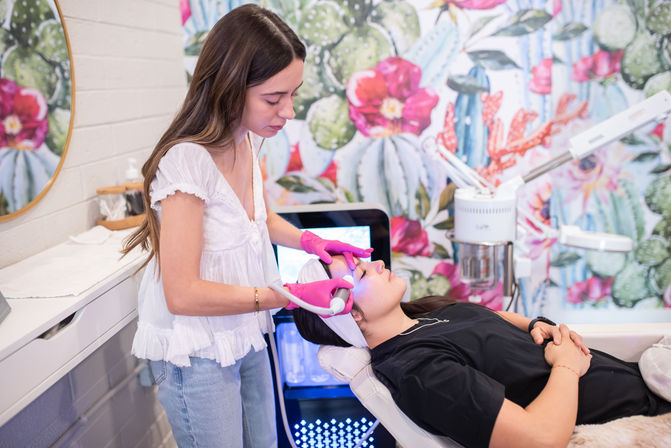 Esthetician wearing pink gloves performing a facial treatment with a handheld device on a client reclining in a spa chair, colorful cactus-and-floral mural and skincare equipment in the treatment room.