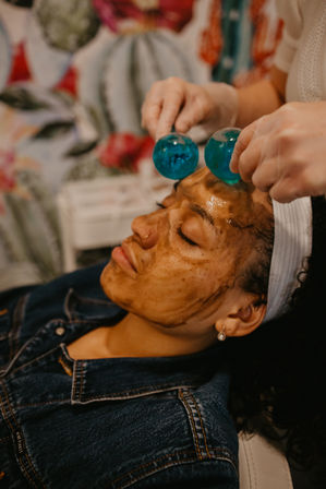 Spa facial treatment: client with a dark mask and headband relaxes as an esthetician wearing gloves rolls two blue glass massage globes across the forehead, skincare and relaxation setting.